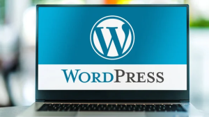 A laptop with a screen displaying the WordPress logo sits in an office or workspace. The background is a gradient blue with a large white "W" in a circle at the top, and the word "WordPress" written below in black and blue text. It's ready for your next website security check.